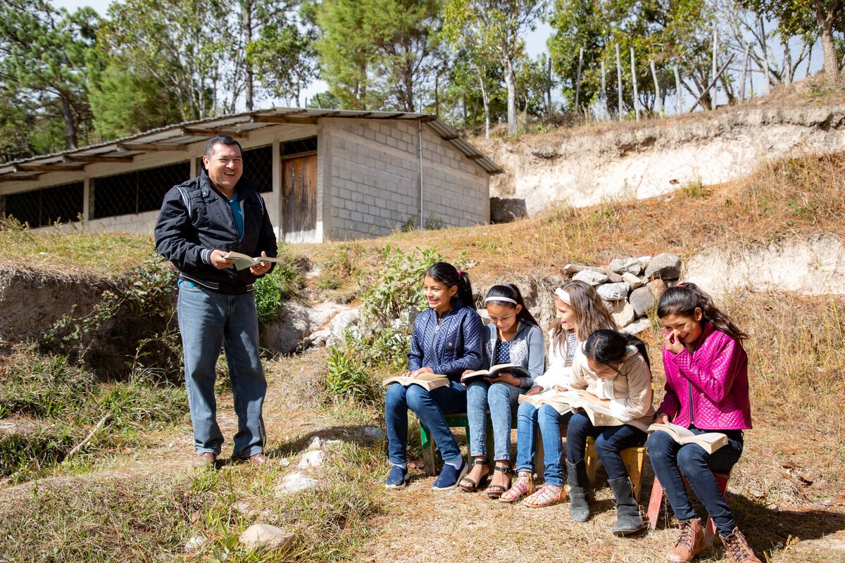 alt="Pastor Genaro in Honduras mit Patenkindern vor der Gemeinde. Kinder lesen Bibel"